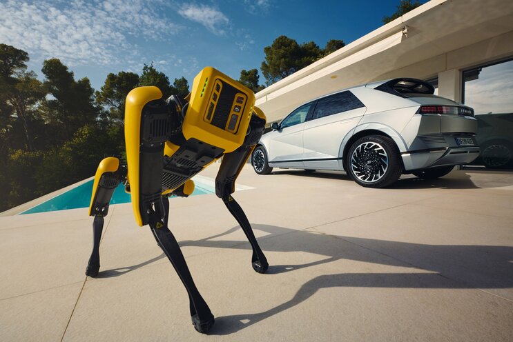 A yellow quadrupedal robot from Boston Dynamics and a silver Hyundai Ioniq 5 car are on a sunlit patio near a pool with trees in the background.