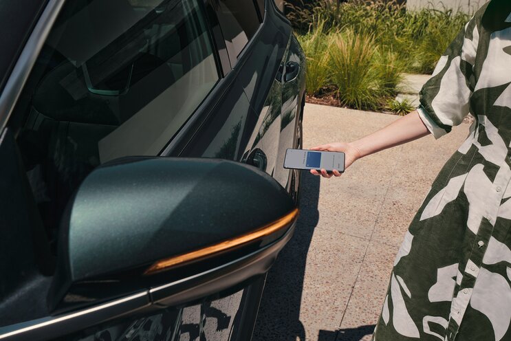 Woman using the Hyundai smartphone app near a car's door to unlock it. Sunlit, outdoor setting with greenery in the background.