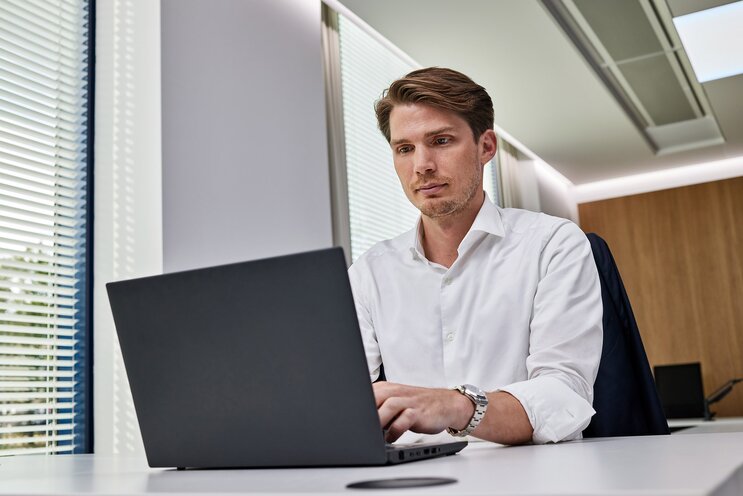 A Hyundai employee seated at a desk in the Hyundai office, wearing a white shirt, and working on a laptop. Blinds covering windows are visible in the background.