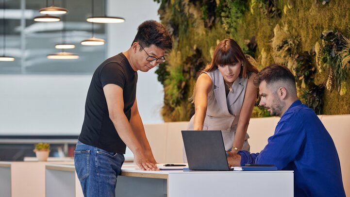 Three people stand and sit around a table in the Hyundai office, engaging in a discussion while looking at a laptop. A green wall with plants decorates the background.