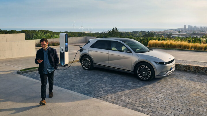 A person walks away from a gray electric Hyundai Ioniq 5 car plugged into a charging station, with wind turbines and a cityscape visible in the background.