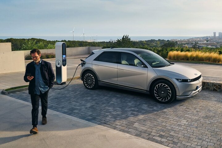 A person walks away from a gray electric Hyundai Ioniq 5 car plugged into a charging station, with wind turbines and a cityscape visible in the background.