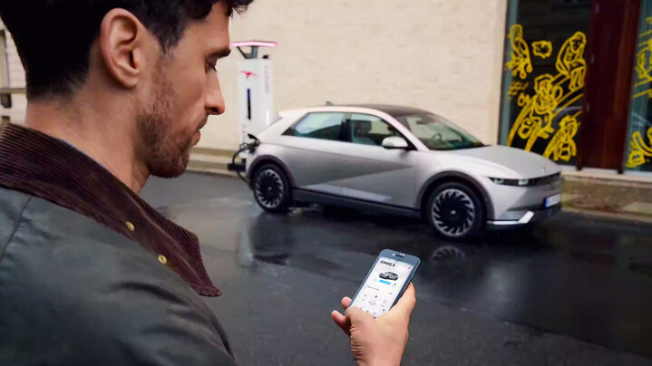 A man using the bluelink smartphone app while standing near a silver Hyundai Ioniq 5 parked on a wet street.