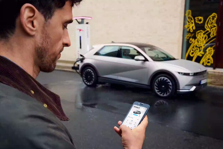 A man using the bluelink smartphone app while standing near a silver Hyundai Ioniq 5 parked on a wet street.