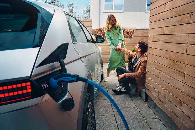 A man kneels and holds hands with a standing woman beside a Hyundai Ioniq 5 charging in a residential area.