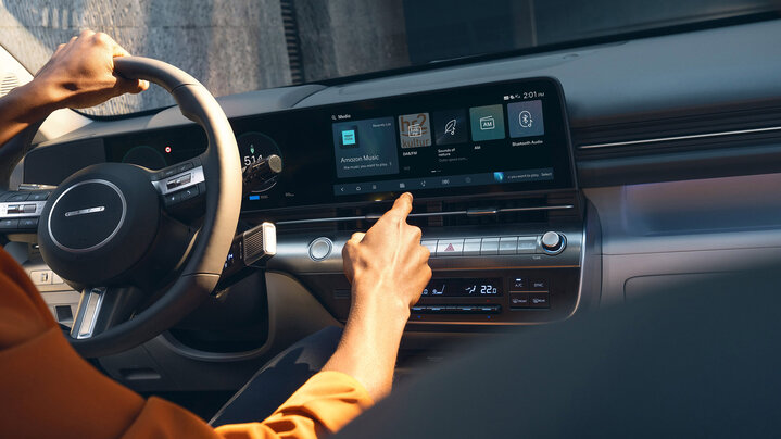 A person’s hand reaches to touch the touchscreen display in a hyundai car interior, with various multimedia options visible on the screen. The other hand is on the steering wheel.