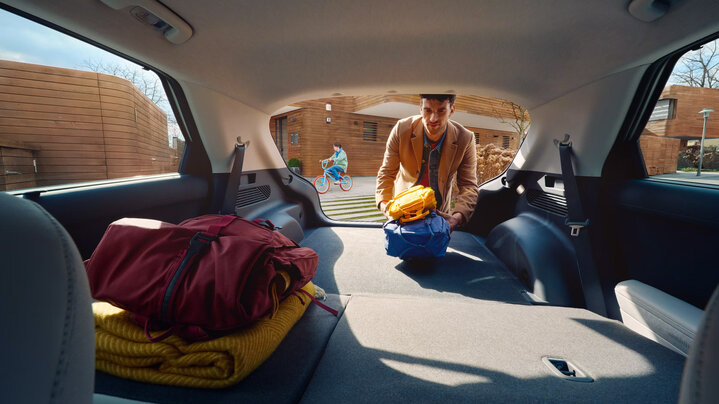 A man places multiple backpacks into the trunk of a Hyundai car with bags already inside, while a child on a bike rides past in the background on a sunny day.