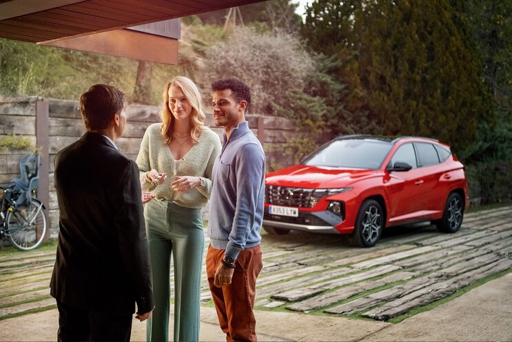 Three people stand conversing next to a parked red Hyundai SUV in a driveway surrounded by greenery. A bicycle is partially visible on the left side.