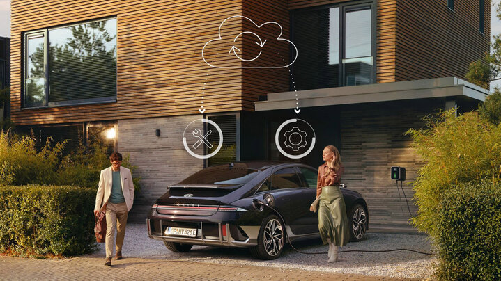 A man walks near a parked electric Hyundai Ioniq 6 car charging outside a modern house, while a woman walks towards him from beside the car. Icons of tools and a cogwheel are shown above the car with dotted lines connecting to a cloud icon.