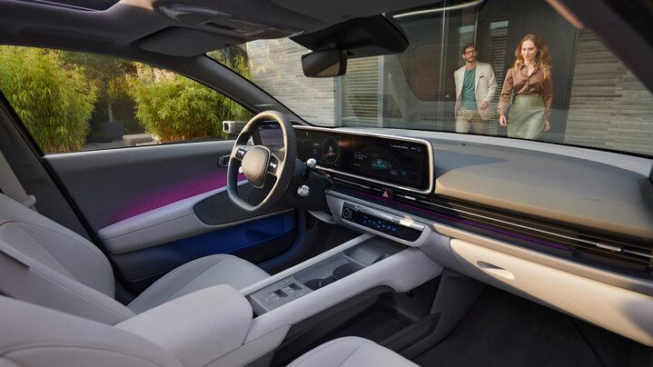 Interior view of a Hyundai Ioniq 6 car with a large touchscreen display and digital dashboard; two people walk towards the car outside near the driver's side door.